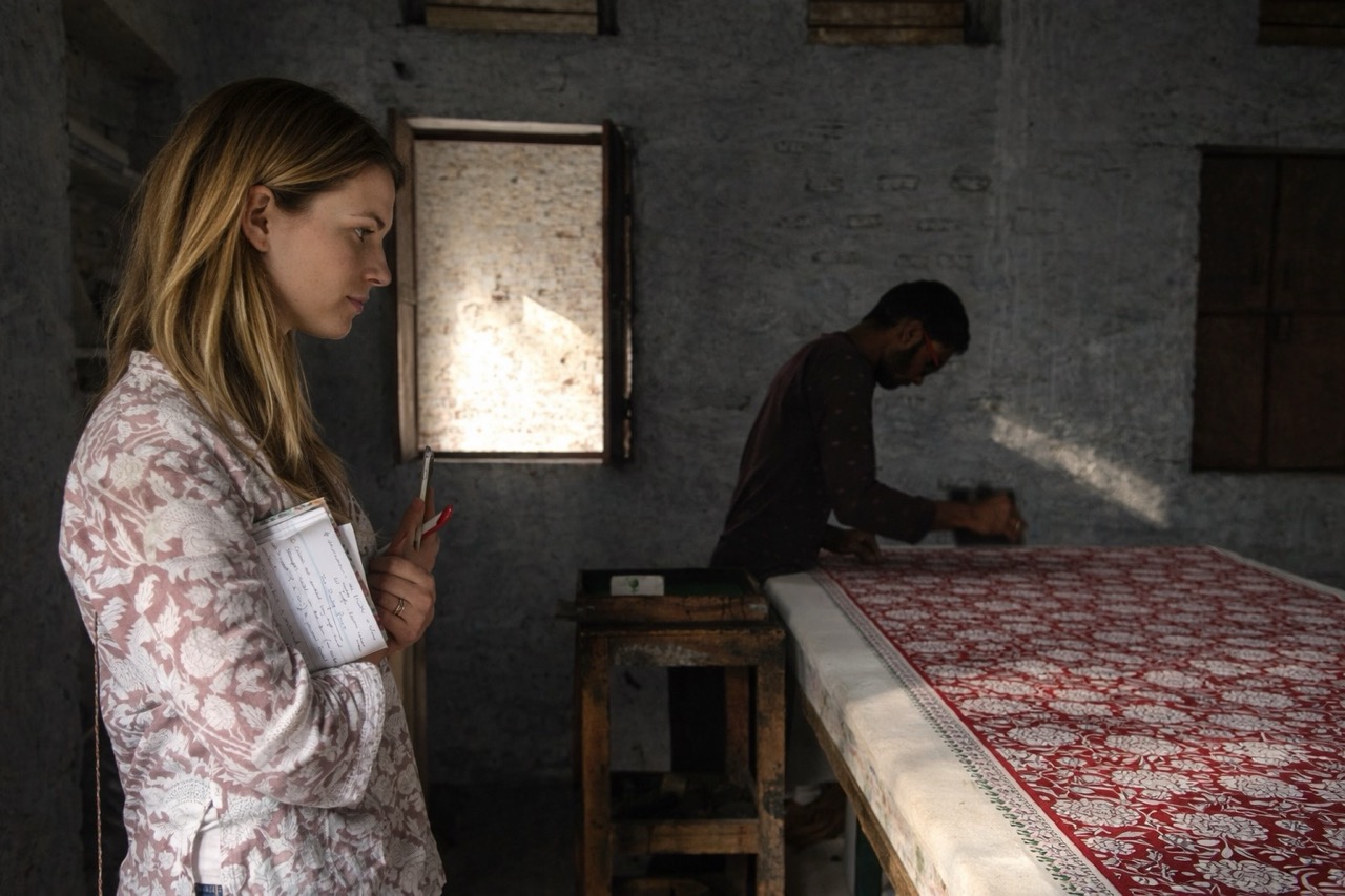 A buyer taking notes beside a printer working a red floral hand block print along a long printing table in a Rajasthan workshop